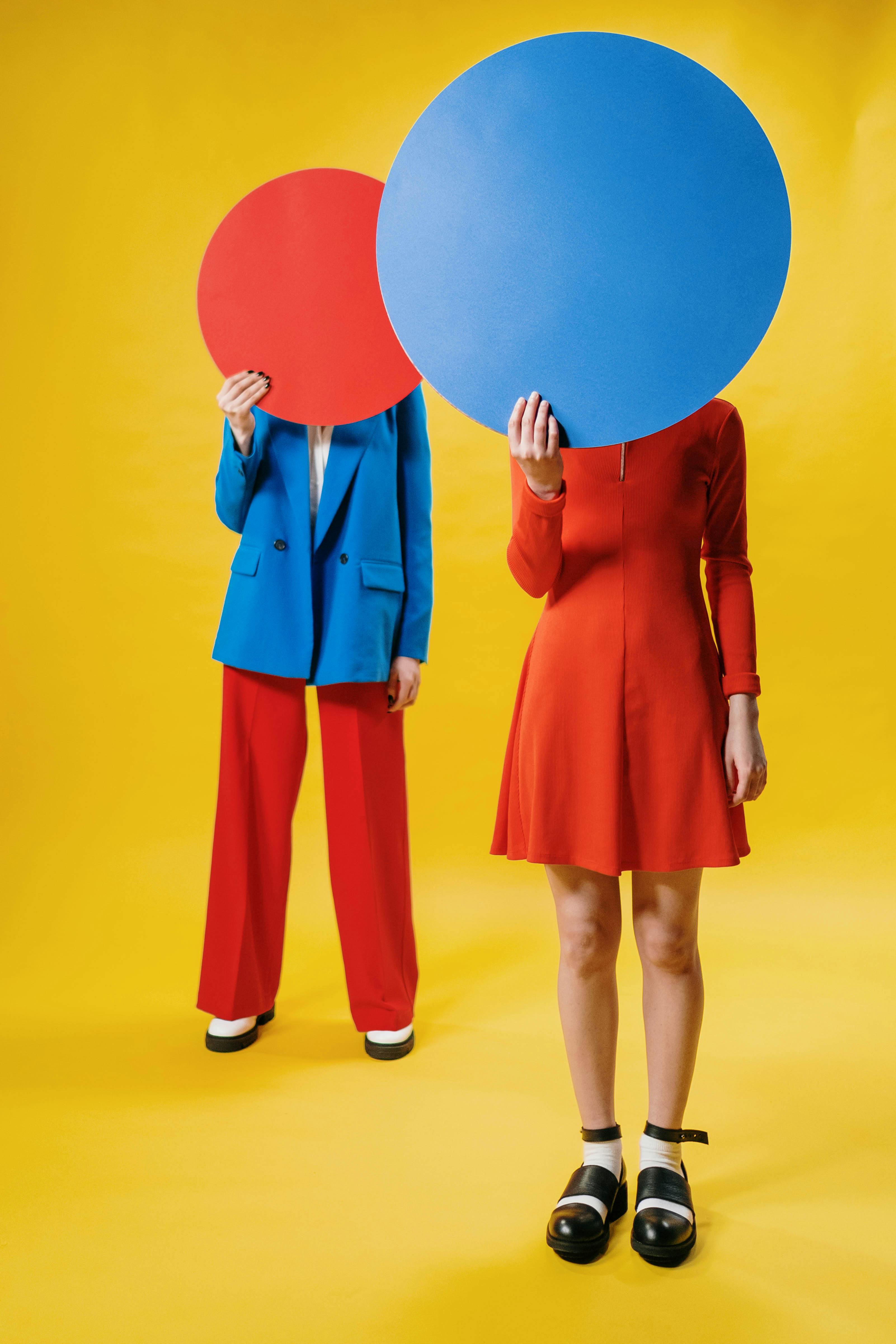 Two figures standing against a yellow background wearing saturated red and blue outfits, faces obscured by circular color blocks.