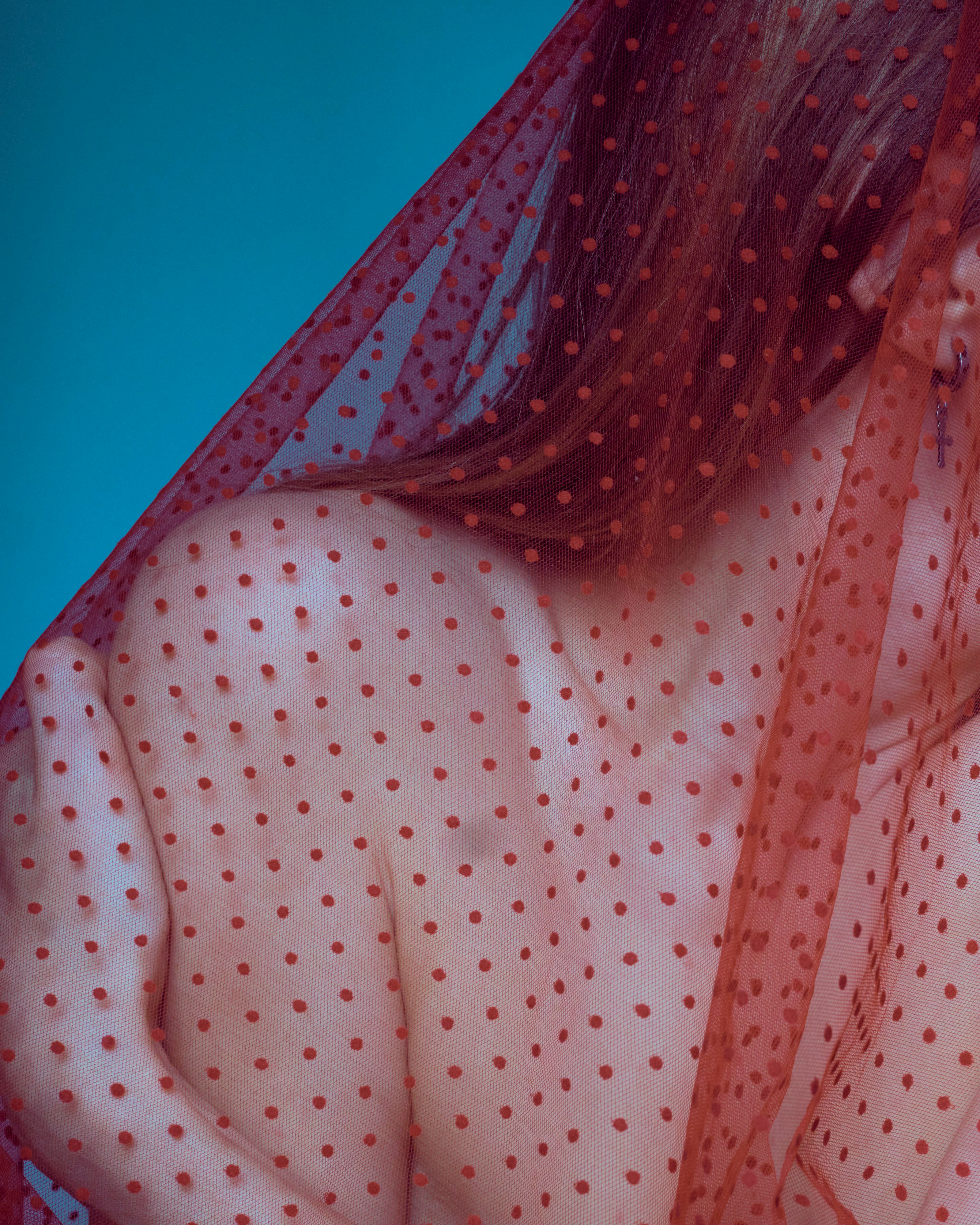 Close-up of a person’s shoulder under a sheer red polka-dot fabric, revealing skin and freckles beneath the veil.